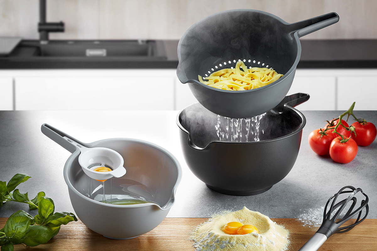 Set aus drei Küchenschüsseln, einem Nudelsieb und einem Ei-Trenner auf einer Küchenarbeitsplatte, umgeben von Tomaten, Eiern und einem Schneebesen