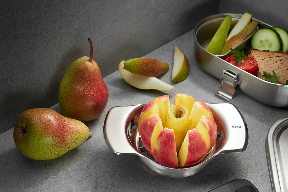 Ein Apfelschneider schneidet einen roten Apfel, daneben liegen frische Birnen und eine Lunchbox mit Obst und Brot