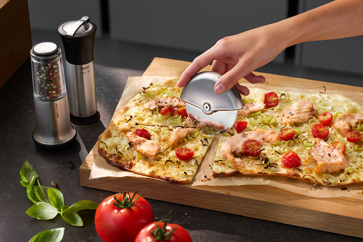 Eine Hand schneidet eine Pizza mit Lachs und Tomaten mit einem Edelstahl-Pizzaschneider. Im Hintergrund befinden sich Pfeffer- und Salzstreuer sowie frische Tomaten und Basilikum auf einem Tisch.