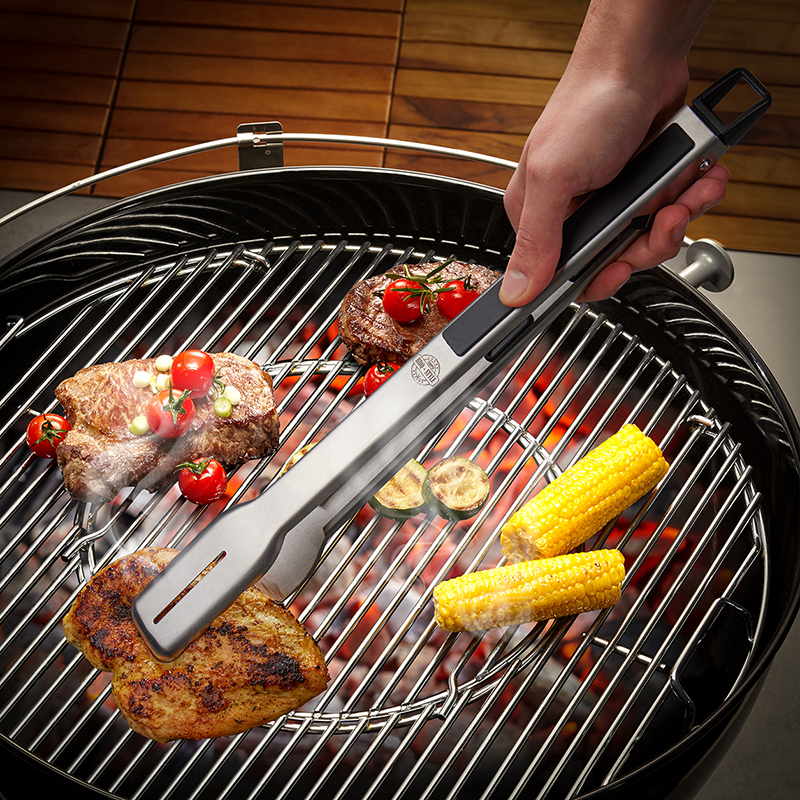 Praktische Grillzange beim Umwenden von saftigem Grillgut auf einem Holzkohlegrill. Eine Hand hält eine Grillzange über einem Holzkohlegrill mit verschiedenen Grillspeisen wie Steak, Hähnchen und Maiskolben.