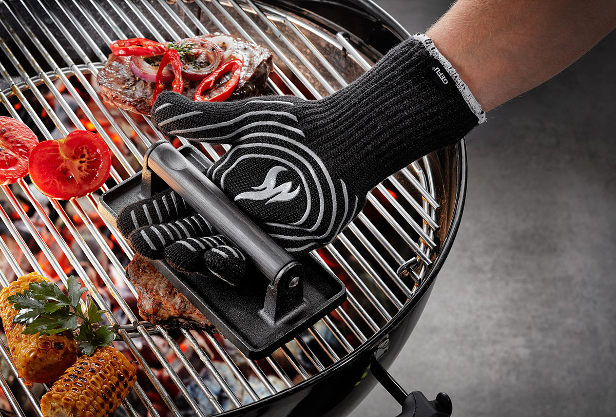 Grillhandschuhe mit Grillpressen-Set auf Holzkohlegrill Eine Person mit Grillhandschuhen verwendet eine Grillpresse auf einem Grill mit Tomaten, Mais und Fleisch.