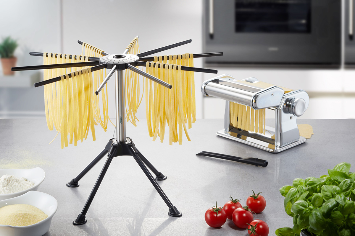 Pasta-Trockengestell und Nudelmaschine in einer modernen Küche Ein Pasta-Trockengestell und eine Nudelmaschine auf einer Küchenarbeitsplatte, umgeben von frischem Basilikum, Tomaten und Zutaten für die Pastazubereitung.