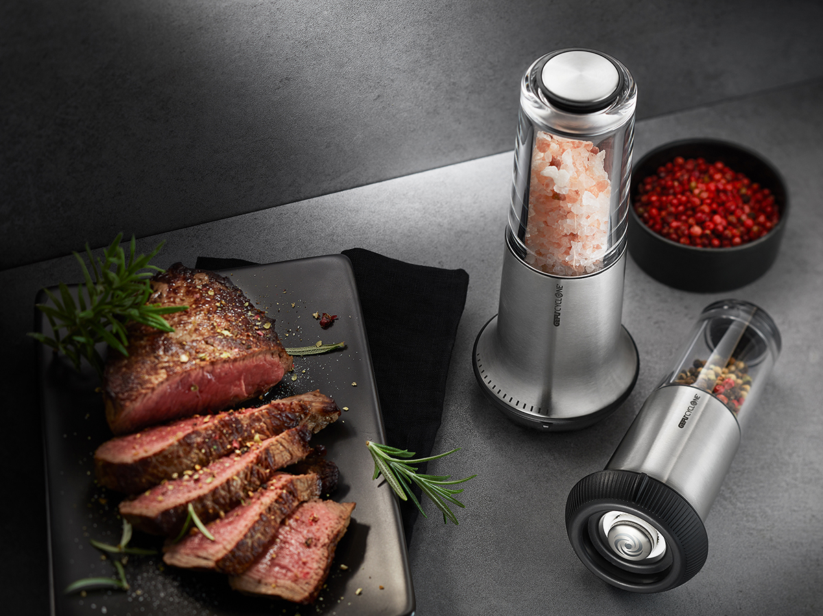 Edelstahl-Salzmühle und Pfeffermühle-Set mit Steakgericht Modernes Set aus Salzmühle und Pfeffermühle aus Edelstahl, präsentiert neben einem perfekt gegarten Steak auf schwarzem Teller.