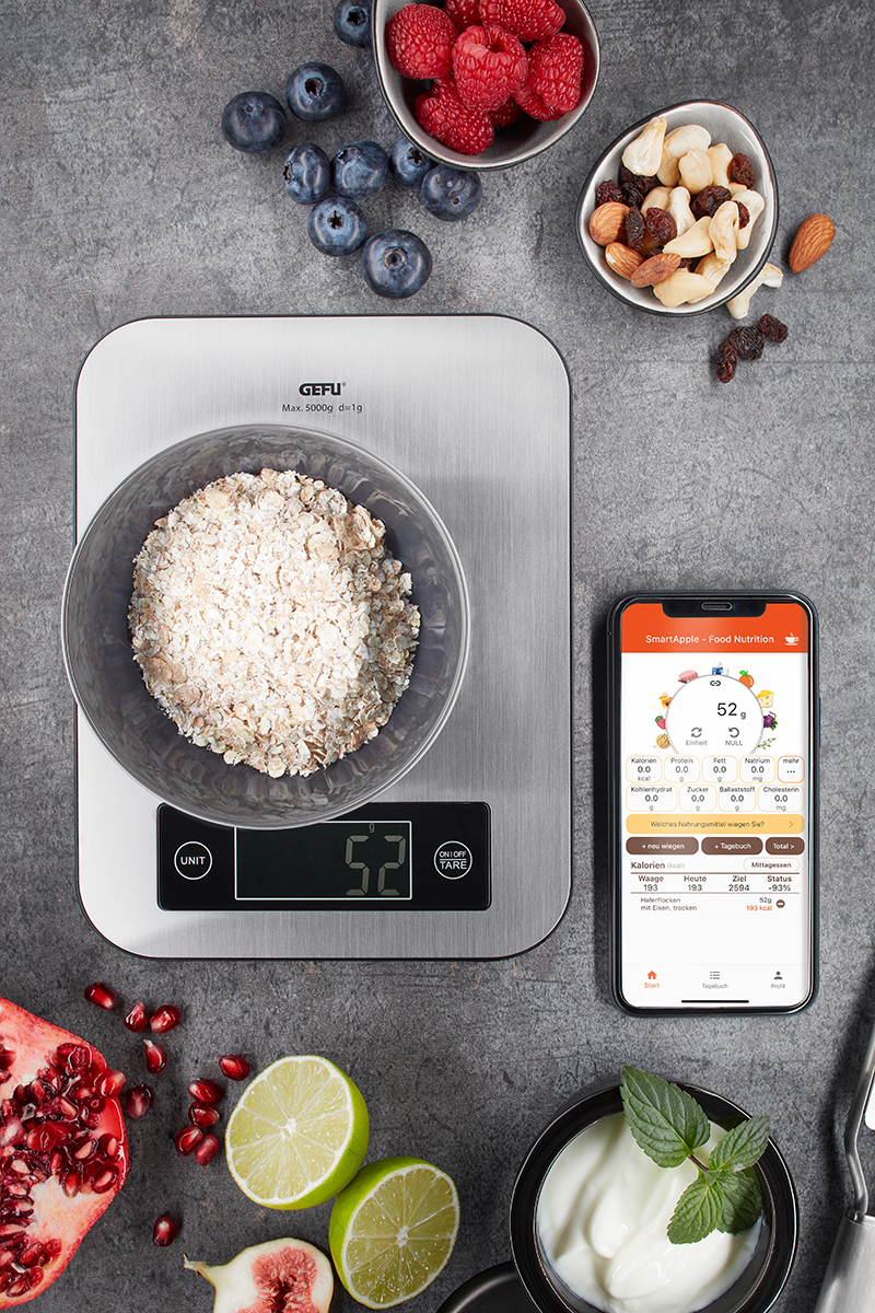 Digitale Küchenwaage mit Smart-App-Steuerung und gesunden Zutaten Digitale Küchenwaage zeigt 52 Gramm Haferflocken, umgeben von Beeren, Nüssen, Limette, Granatapfel und Smartphone mit Ernährungs-App
