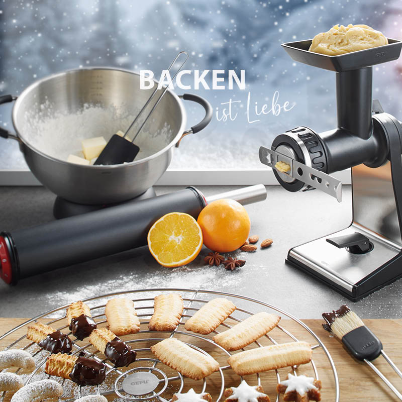 Eine Küche mit Backzubehör, Schüssel, Teigrolle, orangene Zitrusfrüchte und Plätzchen auf einem Gitter mit dem Schriftzug 'Backen ist Liebe'.
