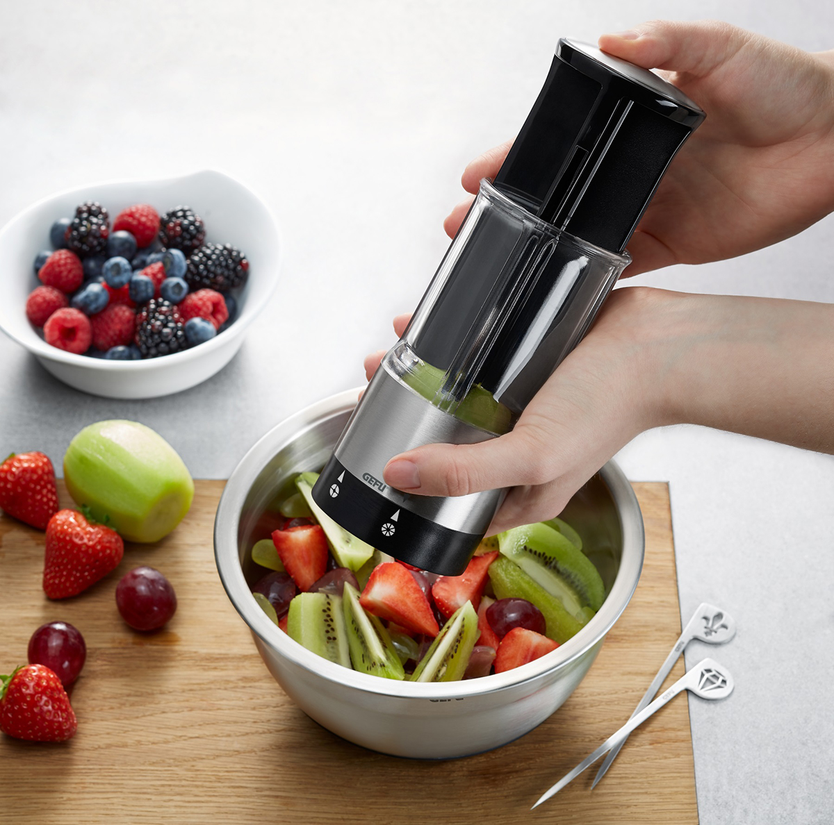 Obstschneider in Aktion zur Zubereitung eines frischen Obstsalats Eine Person benutzt einen Obstschneider, um Kiwi und Erdbeeren für einen Obstsalat zu schneiden, neben einer Schale mit Beerenfrüchten.