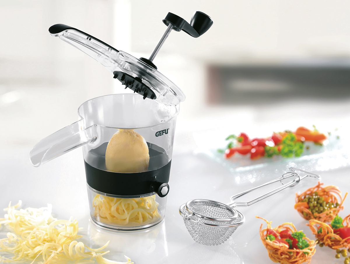 Effizienter Gemüseschneider mit transparentem Behälter und Kurbel für spiralisierte Gemüse Manueller Gemüseschneider aus Kunststoff mit Handkurbel, ideal für das Herstellen von Spiralgemüse, abgebildet mit einer Kartoffel und geschnittenem Gemüse im Hintergrund.