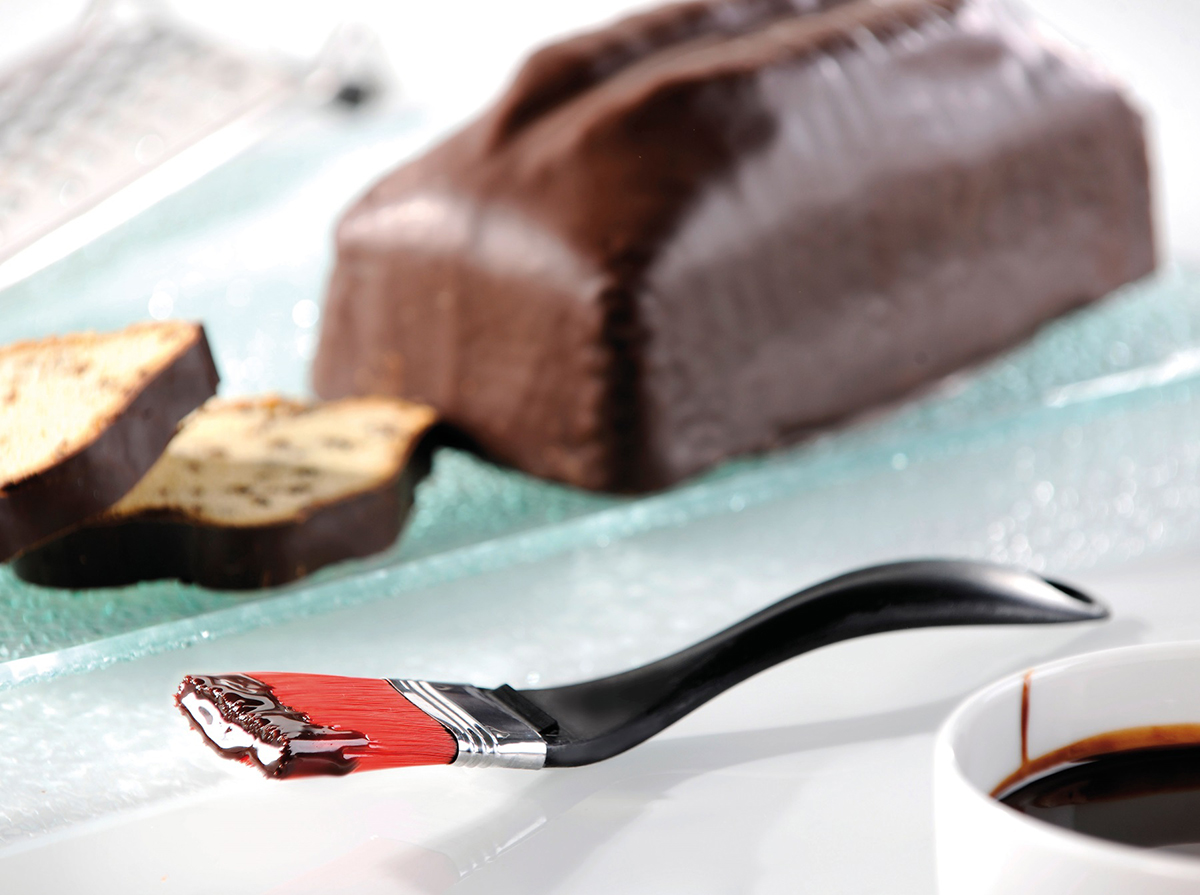 Frisch gebackener Schokoladenkuchen mit Glasur und Backpinsel In der Nahaufnahme ein Schokoladenkuchen mit glänzender Glasur. Ein Pinsel mit dunkler Schokolade und eine Tasse befinden sich im Vordergrund.