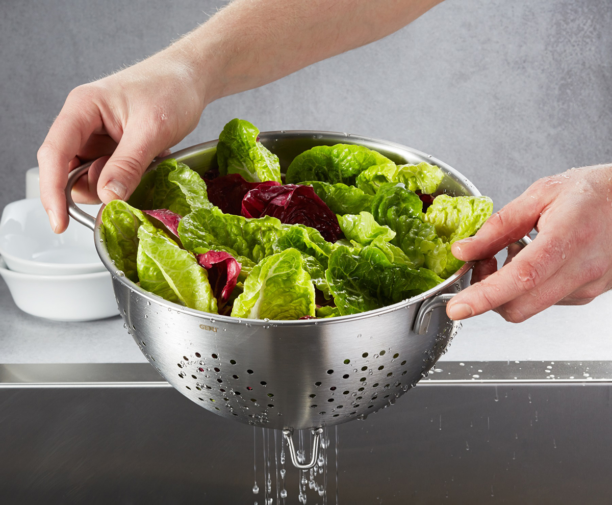 Frischer Salat im Edelstahlsieb über der Spüle Hände halten ein Edelstahlsieb mit frischem Salat, während Wasser abläuft