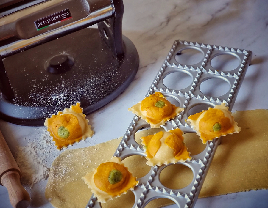 Kürbisravioli Selbstgemachte Ravioli, gefüllt mit Kürbis und Erbsen, liegen auf einem Raviolibrett neben einer Nudelmaschine auf einer bemehlten Oberfläche.