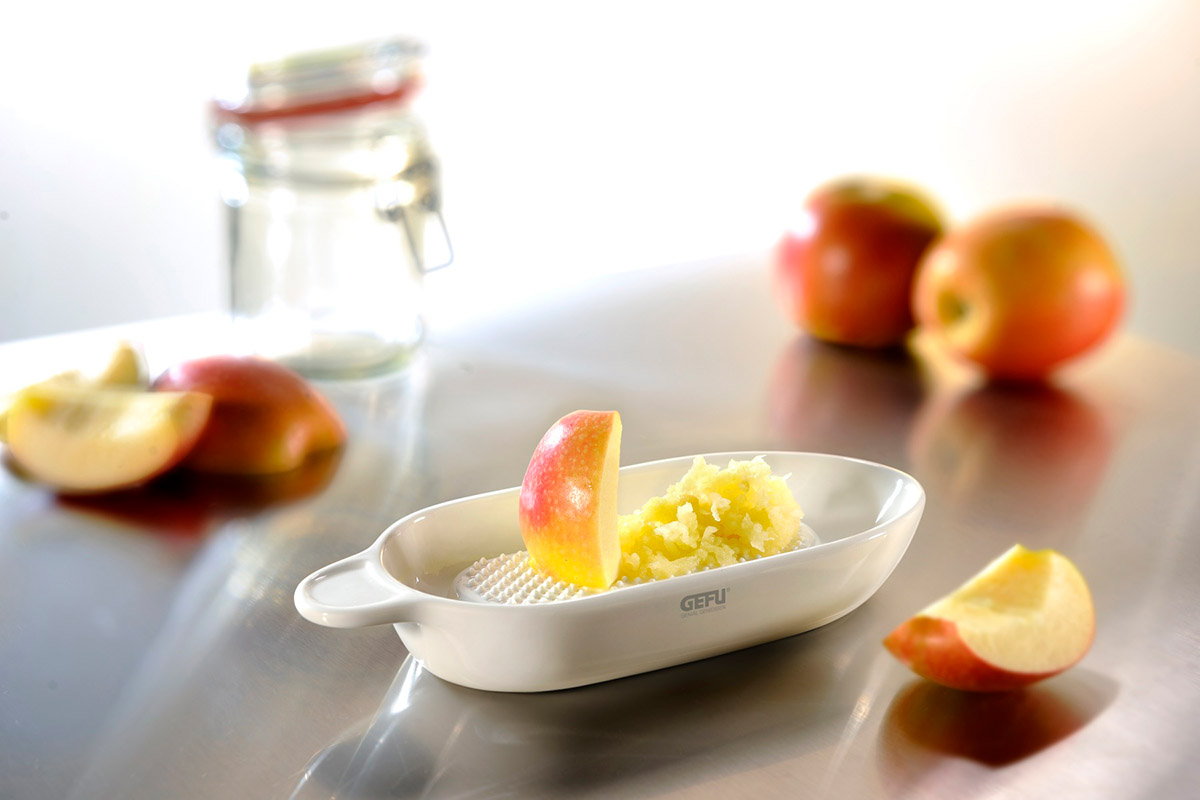 Apfelreibe mit geriebenem Apfel und frischen Apfelscheiben Eine weiße Apfelreibe mit geriebenem Apfel, daneben frische Apfelscheiben und Äpfel im Hintergrund