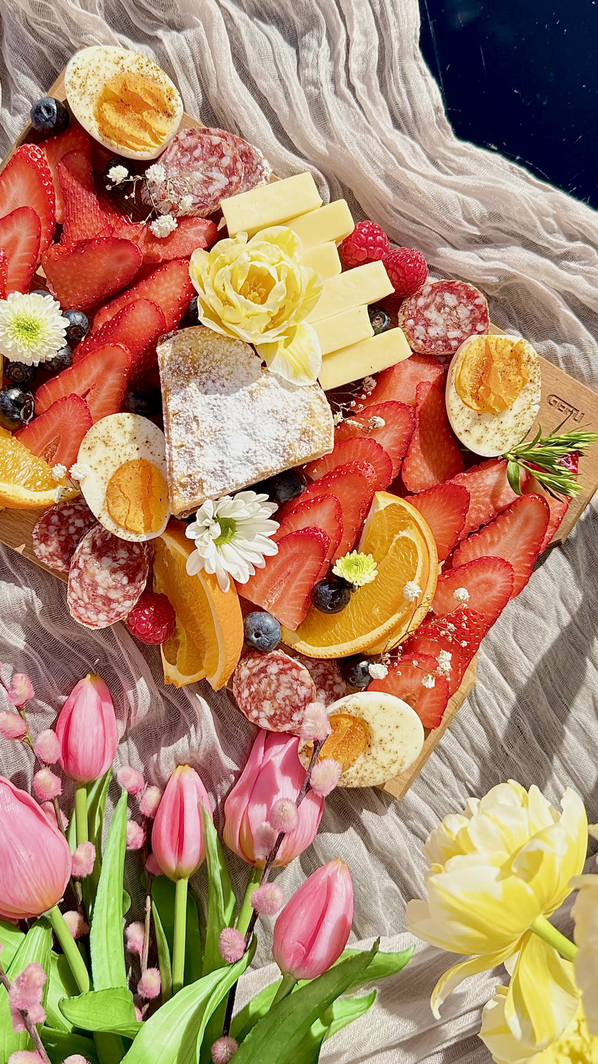 Frühlingsbrunch  Eine dekorative Platte mit Erdbeeren, Orangen, Käse, Wurst, einem gekochten Ei und Blüten, umgeben von pinken Tulpen und gelben Blumen auf einem Tuch.