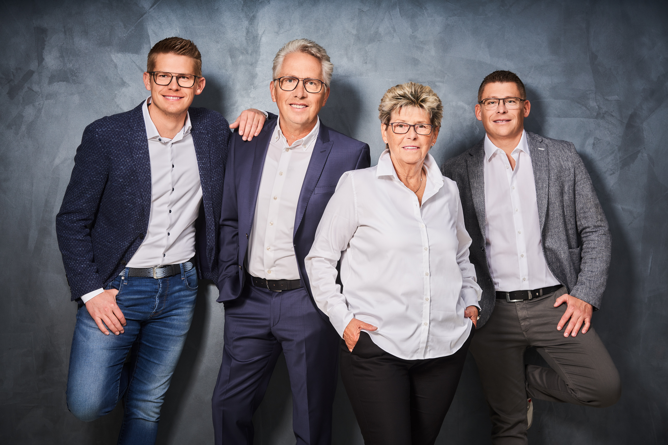 Geschäftsteam posiert lächelnd vor grauem Hintergrund Vier Personen in Business-Outfits stehen gemeinsam vor einem grauen Hintergrund, lächelnd in die Kamera blickend.