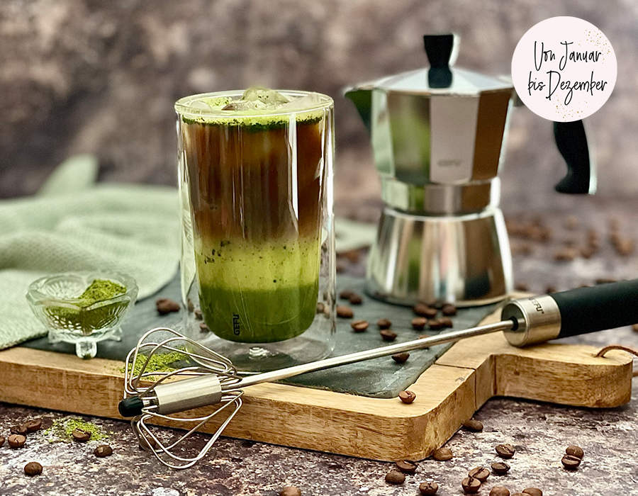 Matcha Espresso Latte  Ein Glas mit Iced Matcha Latte, einer Kaffee-Maschine und Zubehör auf einem Holztablett, umgeben von Kaffeebohnen und einem Schneebesen.