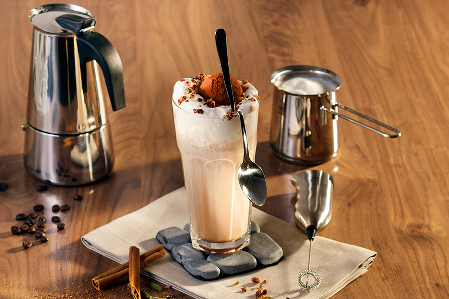 Iced coffee Oriental Ein Glas mit Eiskaffee, Sahne und Schokoraspeln, daneben ein Espressokocher, Milchaufschäumer und Kaffeebohnen auf einem Tisch