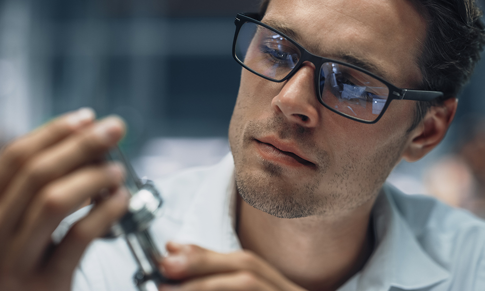 Mann mit Brille inspiziert ein technisches Bauteil Ein Mann mit Brille hält ein technisches Bauteil und inspiziert es genau, konzentriert auf die Arbeit.