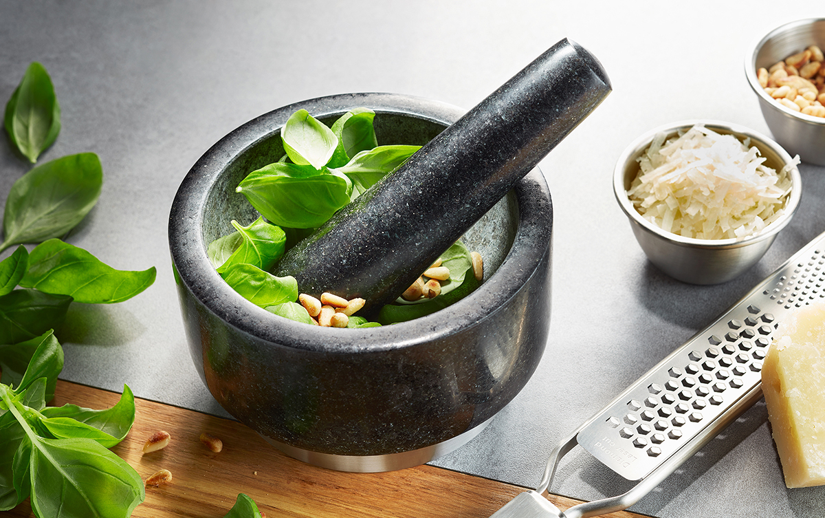 Mörser mit frischem Basilikum, Pinienkernen und Käsereibe Ein Mörser gefüllt mit frischem Basilikum und Pinienkernen, daneben geriebener Käse und eine Käsereibe auf einem Küchentisch