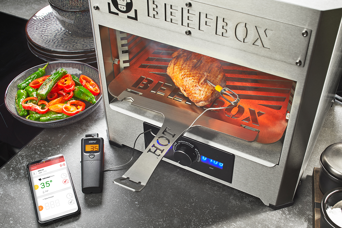 Ein Beefbox Grill mit einem gegrillten Stück Fleisch, umgeben von einem Smartphone zur Temperaturüberwachung und einem Thermometer, daneben eine Schale mit frischem Gemüse.