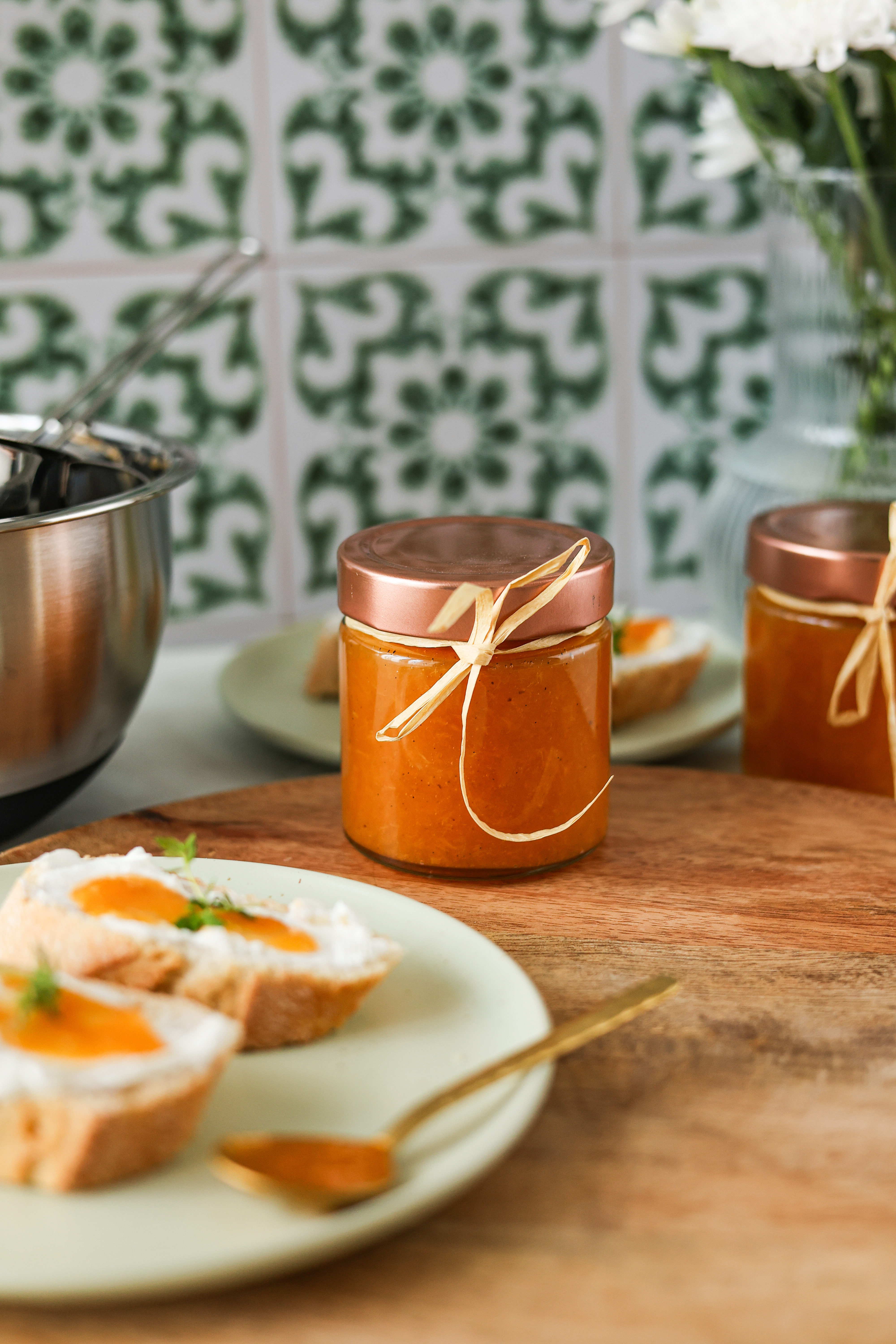 Gewürz-Aprikosen Aufstrich  Ein Glas selbstgemachter Marmelade, dekoriert mit einer Schleife, steht auf einem Holzbrett neben Baguettescheiben und einem Blumenstrauß im Hintergrund.