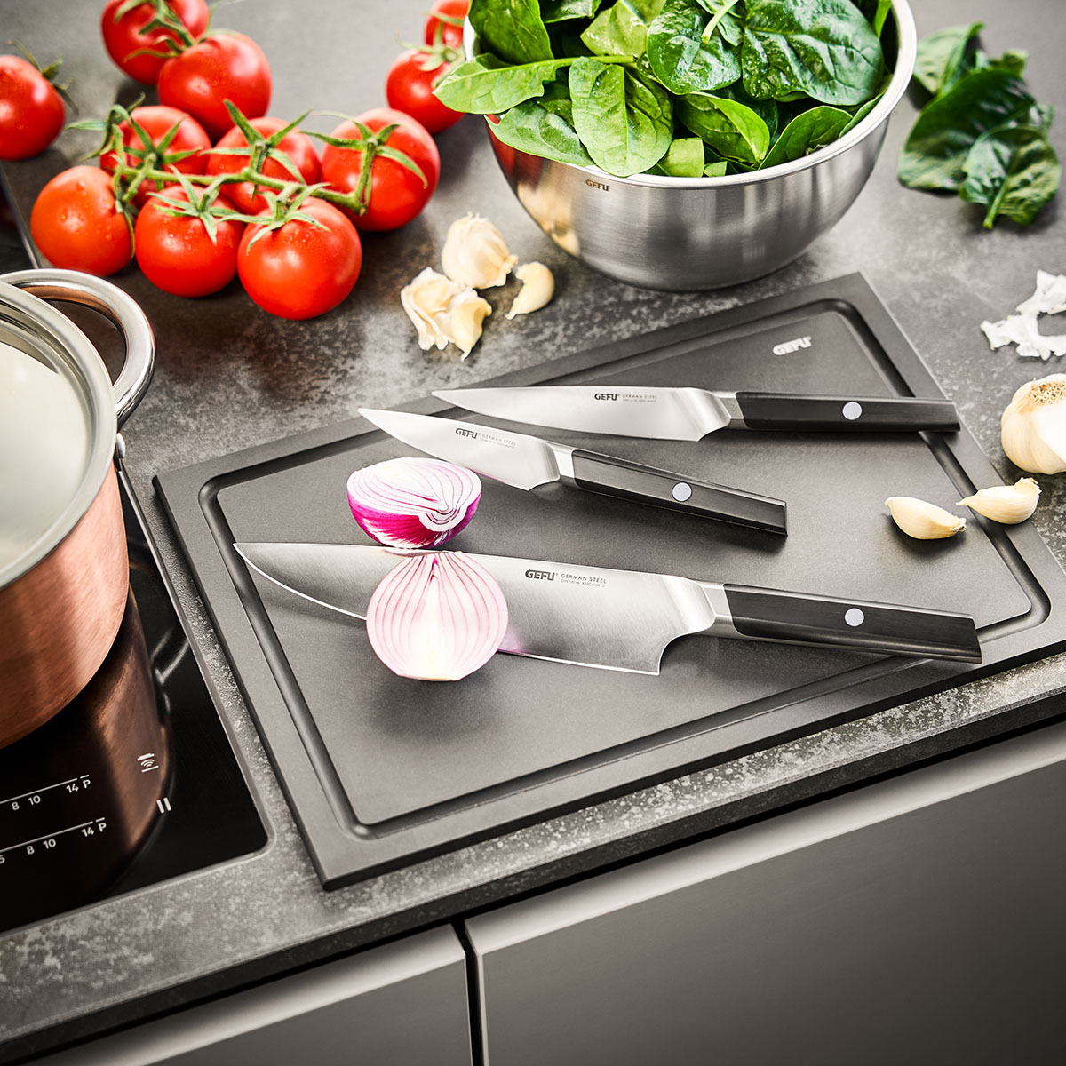 Drei Gefu Messer auf einem schwarzen Schneidebrett, umgeben von frischen Tomaten, Spinatblättern und Zwiebeln in einer modernen Küchenumgebung
