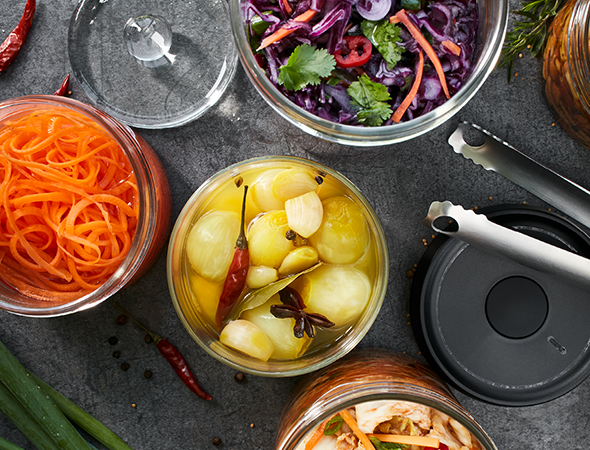 Vielfalt an eingelegtem Gemüse in Glasbehältern auf dunkler Oberfläche Obenansicht von verschiedenen eingelegten Gemüsen in Gläsern, darunter Karotten, Zwiebeln und Rotkohl, auf einer dunklen Tischplatte mit Küchenzubehör