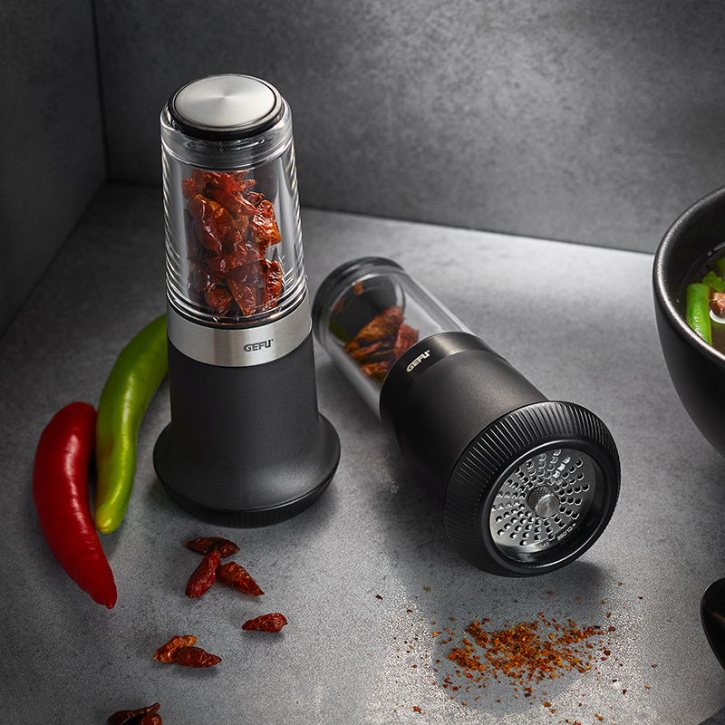 Elegante Gewürzmühlen mit getrockneten Chilischoten Zwei schwarze Gewürzmühlen aus Edelstahl und Glas mit getrockneten Chilis auf einer grauen Oberfläche, daneben frische grüne und rote Chilischoten.