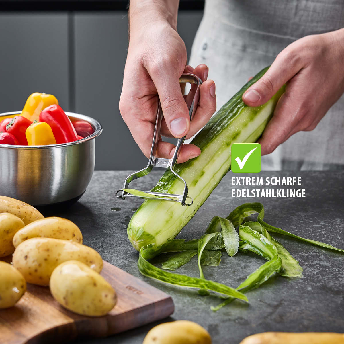 Person verwendet einen Gemüsehobel zum Schälen einer Gurke, daneben Kartoffeln und Paprika in einer Schüssel
