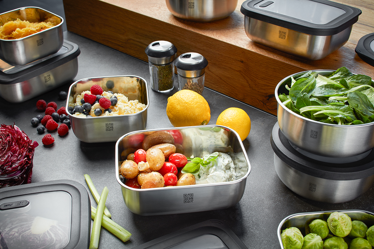 Edelstahl-Lunchboxen mit frischem Salat und Beeren Mehrere Edelstahl-Lunchboxen auf einer Küchentheke, gefüllt mit Salat, Beeren und anderen frischen Zutaten. Daneben liegen Zitronen und Gewürzstreuer.