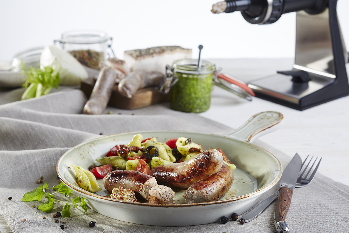 Salsiccia mit mediterranem Pastasalat Eine Pfanne mit gebratener Bratwurst und Nudelsalat, serviert auf einem Leinentuch, im Hintergrund Kräuterpesto und Frühstückszutaten.