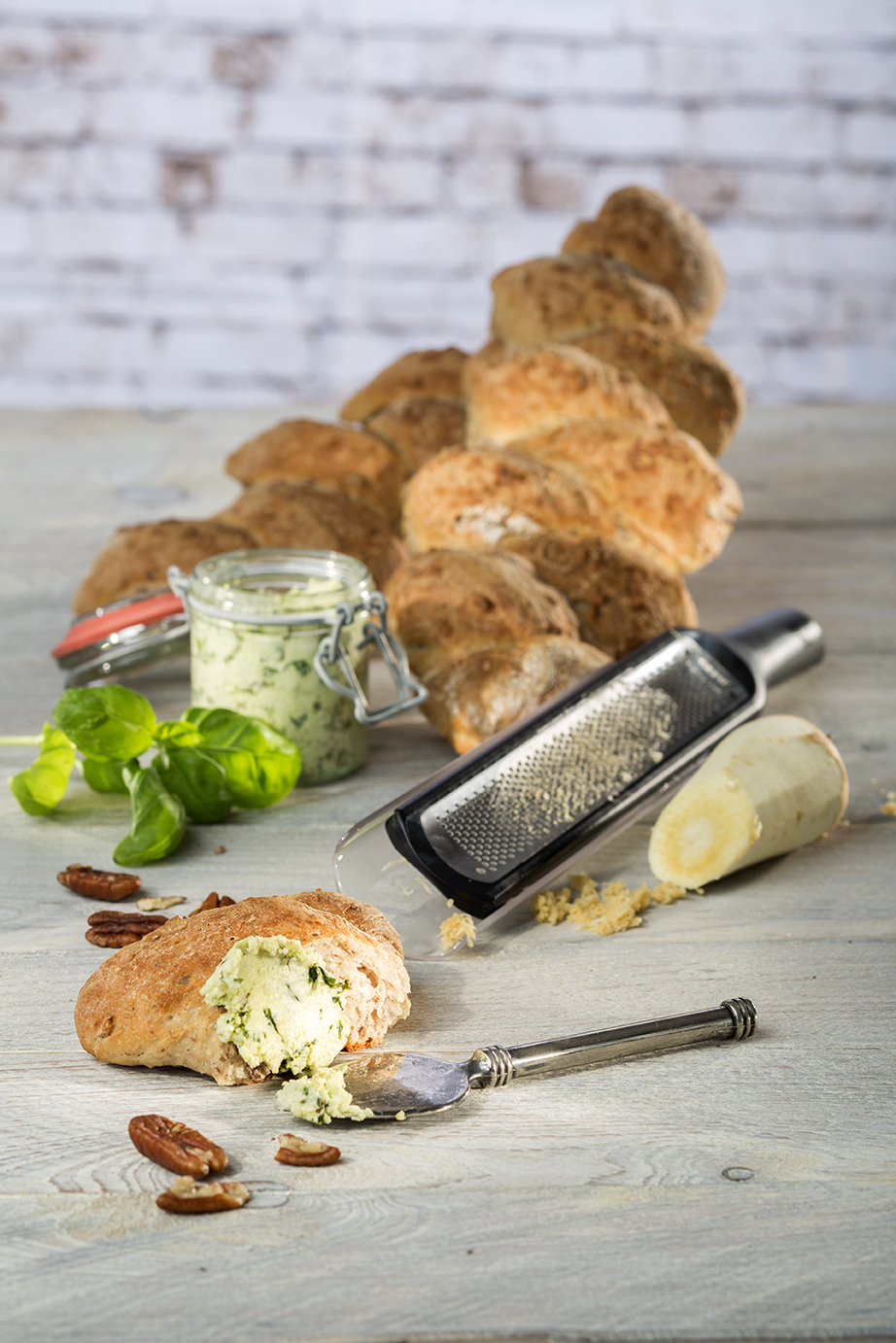 Pastinaken-Ährenbaguette mit Parmesanaufstrich  Ein rustikales Brot mit einem Messer, das mit Kräuterbutter bestrichen ist, ein Glas mit Kräuterbutter und eine Reibe neben frischem Meerrettich und Pekannüssen