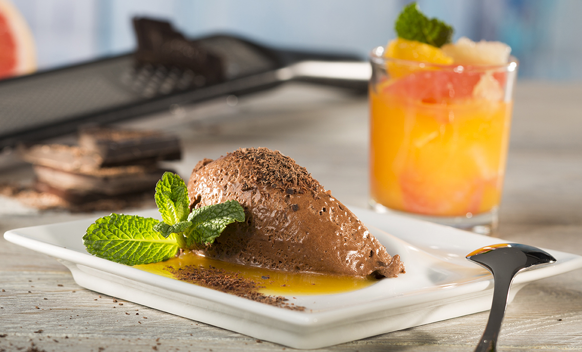 Ginger chocolate mousse with citrus fruit compote  Ein Teller mit reichhaltigem Schokoladenmousse, garniert mit frischen Minzblättern, im Vordergrund; im Hintergrund ein Glas mit einem erfrischenden Frucht-Cocktail.