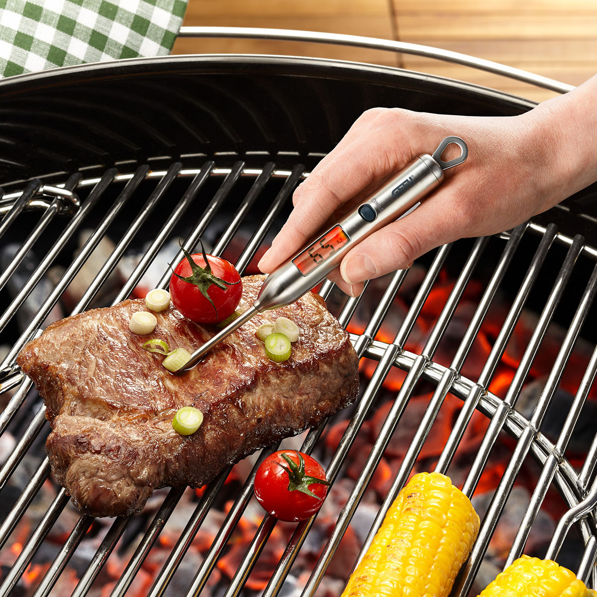 Digitales Grillthermometer mit Steak und Gemüse auf dem Grill Hand verwendet digitales Grillthermometer zur Temperaturmessung eines Steaks neben gegrillten Tomaten und Maiskolben