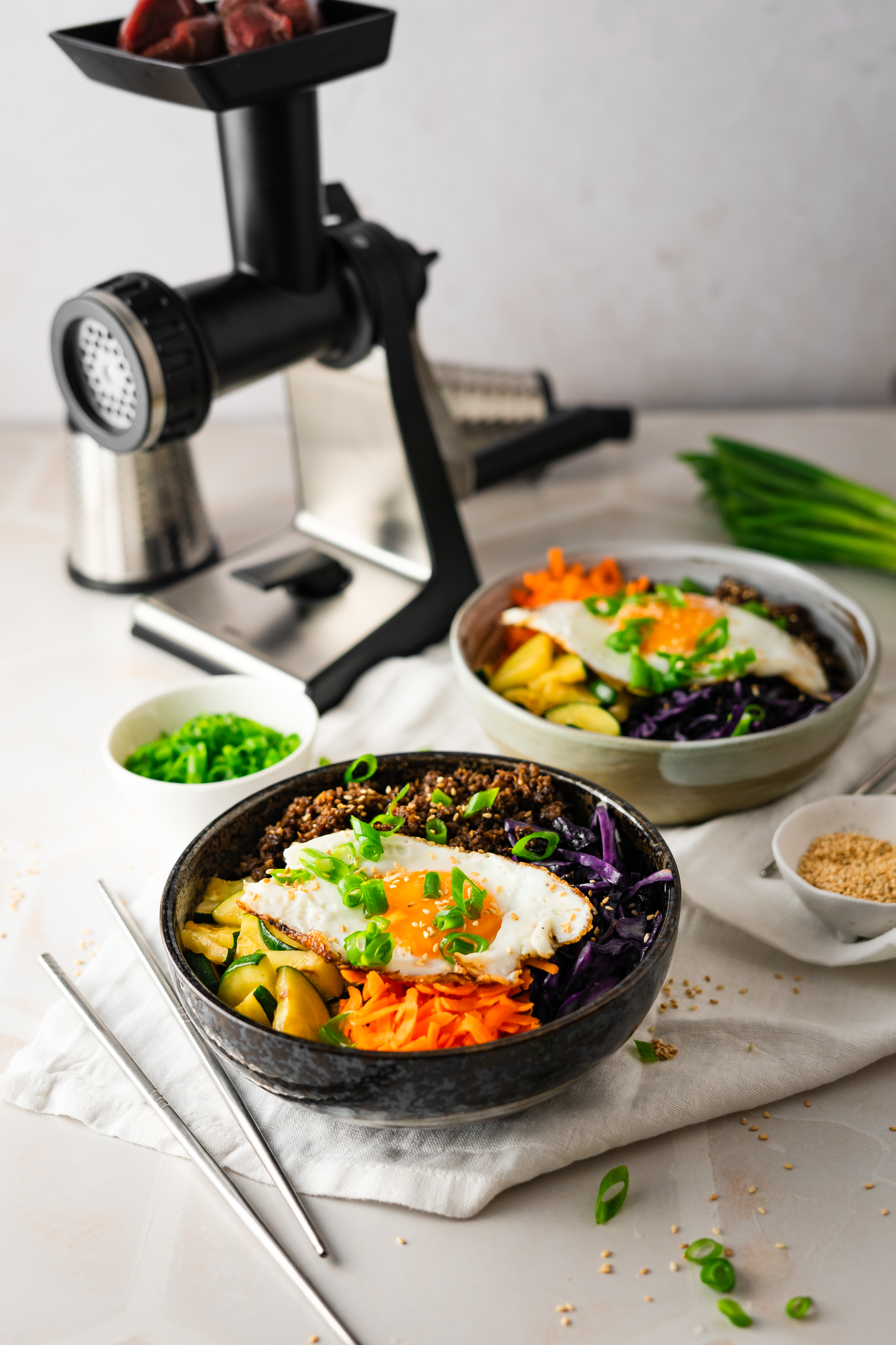 Bulgogi Beef Bowl Bibimbap-Bowl mit Gemüse, Rindfleisch, Spiegelei und frischen Kräutern, serviert neben einem Fleischwolf auf einem Tisch