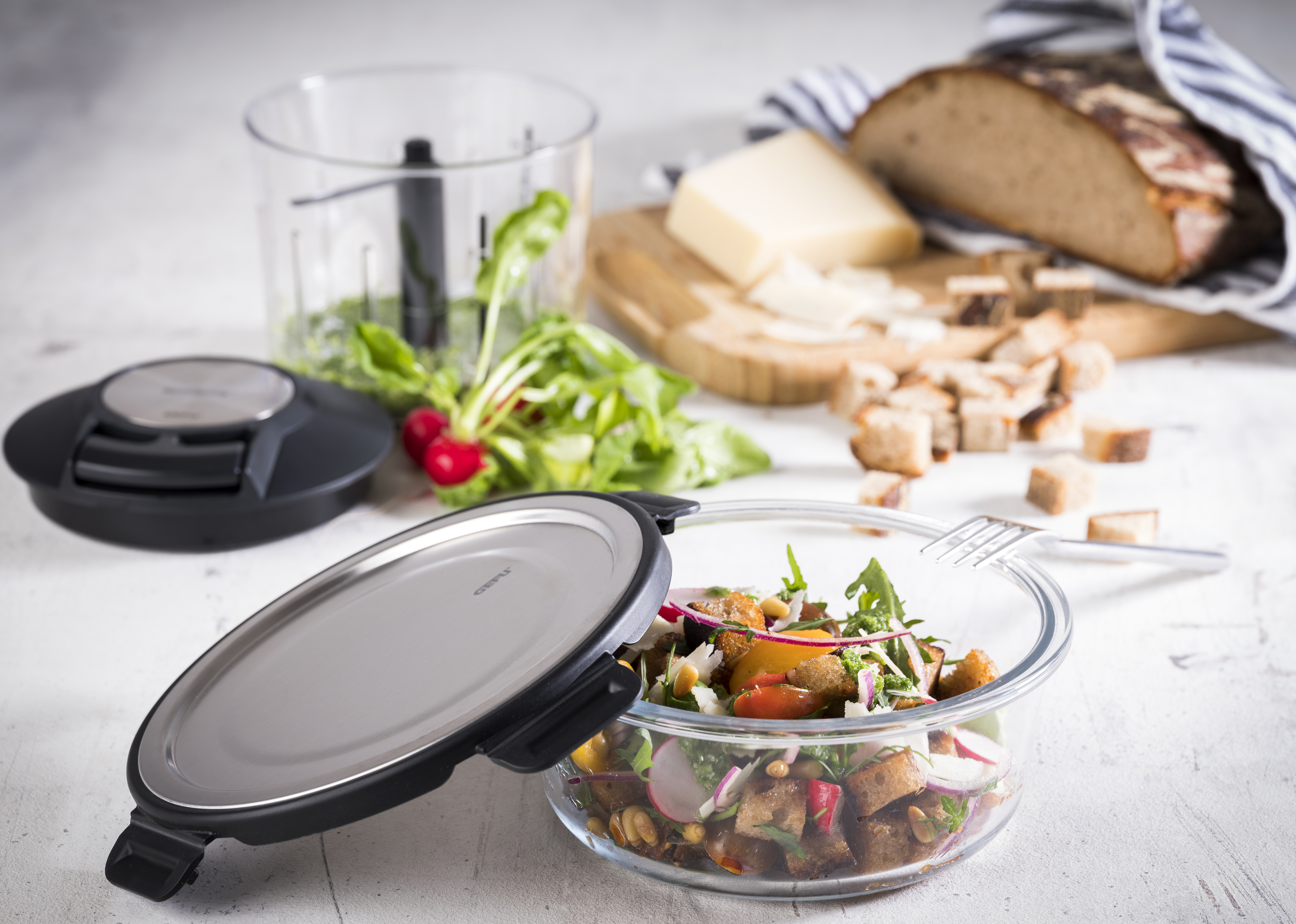 Mediterraner Brotsalat mit Parmesan, Honig und Radieschenblatt-Pesto  Ein Salat mit Gemüse in einer Glasschüssel, umgeben von Küchenutensilien und Brotwürfeln auf einem Tisch.