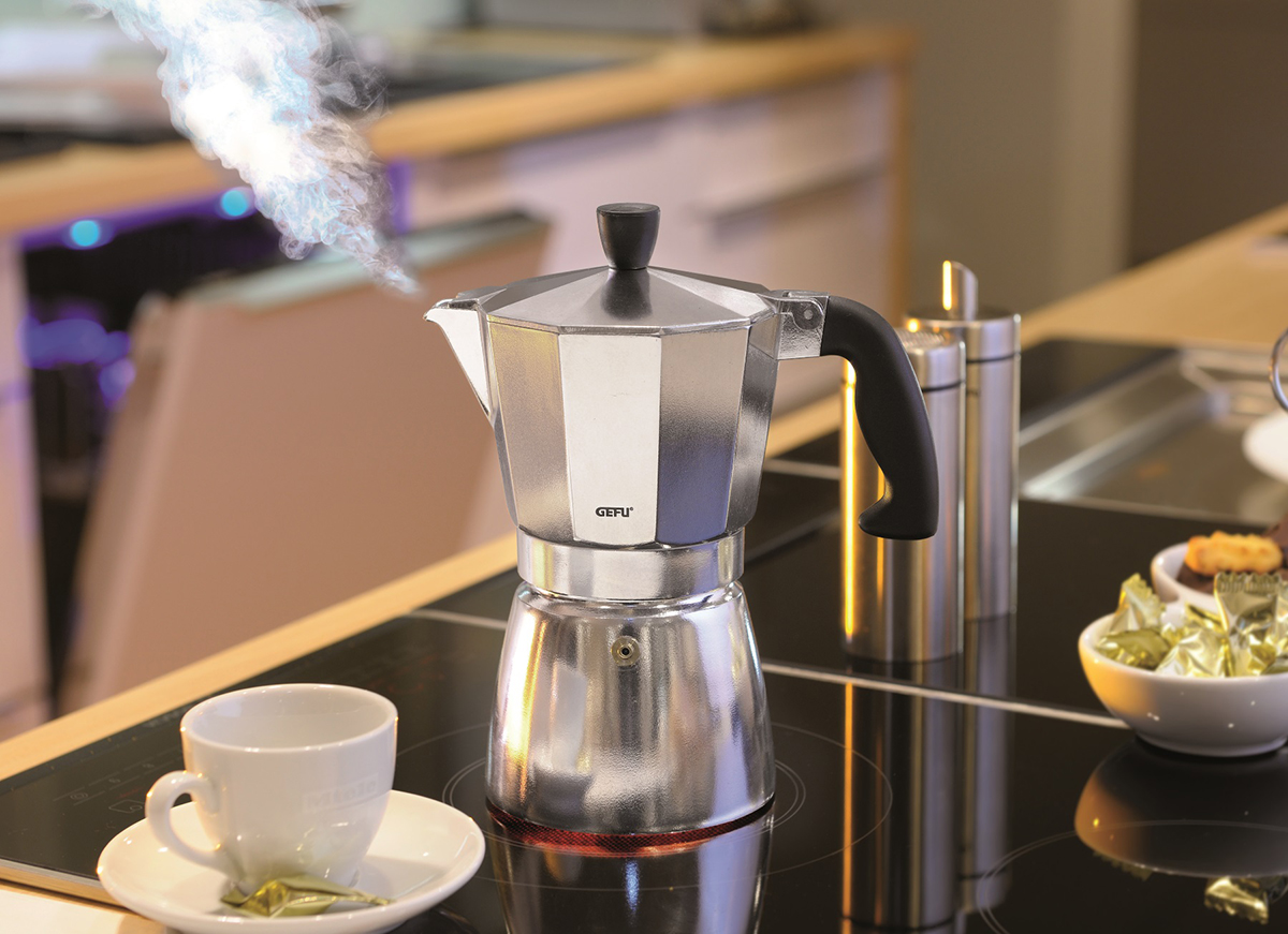 Ein Espressokocher aus Edelstahl steht auf einem modernen Herd und dampft. Daneben steht eine Tasse mit Untertasse.