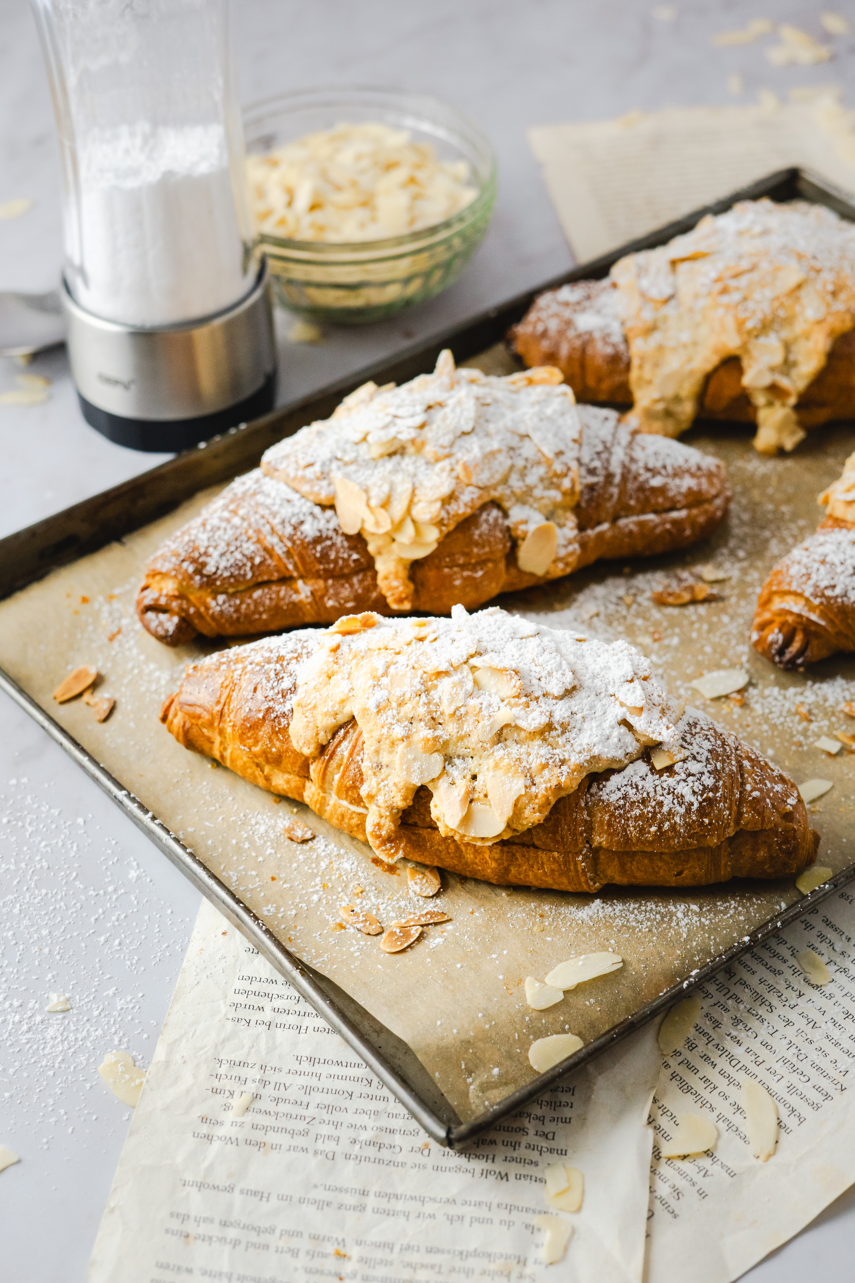 Almond Croissants Nahaufnahme von frischen Croissants mit Mandeln und Puderzucker auf einem Backblech liegend, im Hintergrund Mandelblättchen in einer Schale und ein Streuer.