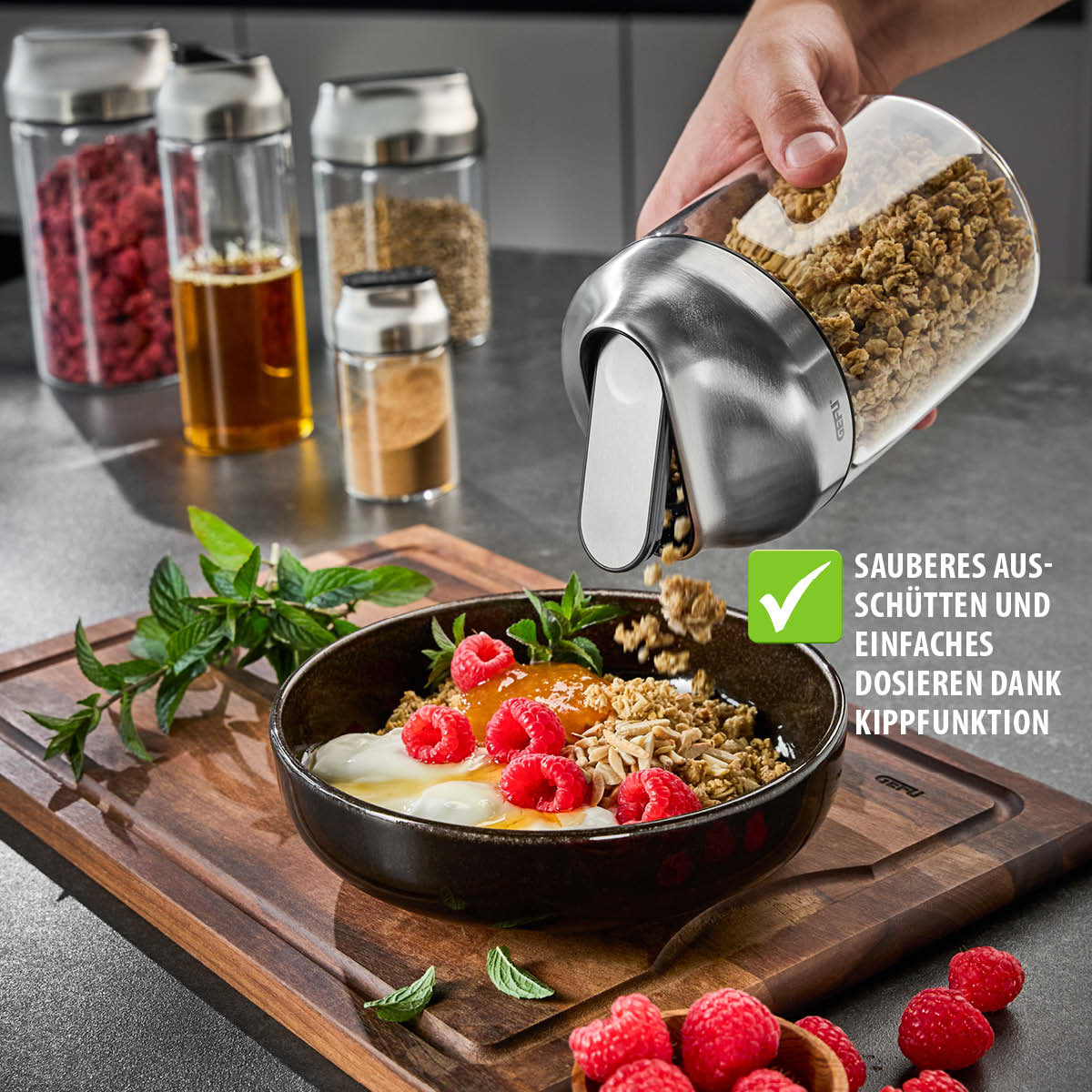 Ergonomischer Glasbehälter mit Kippfunktion für einfaches Dosieren von Müsli Ergonomischer Glasbehälter schüttet Müsli über eine Schüssel mit Joghurt und Früchten, ideal für sauberes und einfaches Dosieren in der Küche