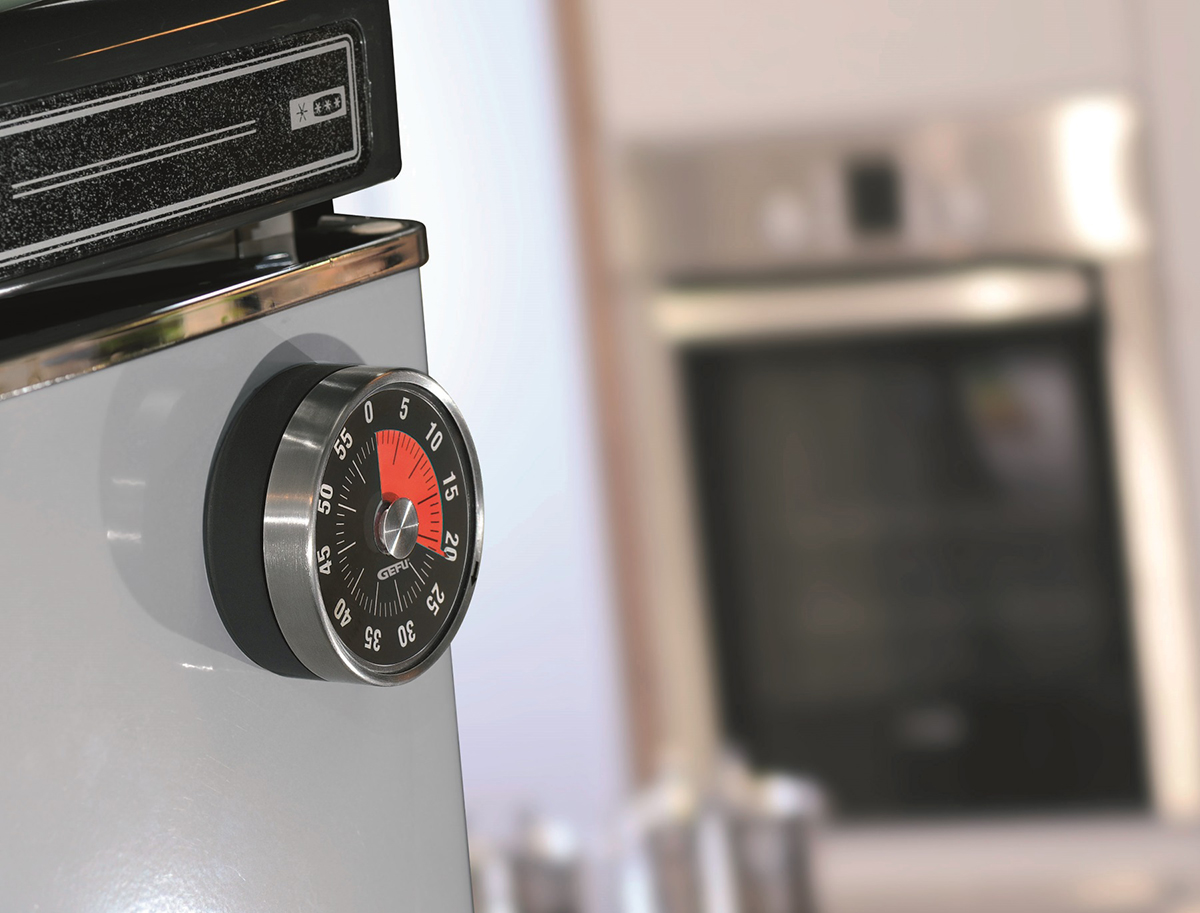 Mechanischer Küchentimer an einem Kühlschrank Ein mechanischer Küchentimer ist an einem Kühlschrank befestigt, im Hintergrund ein unscharfer Blick auf einen Ofen.