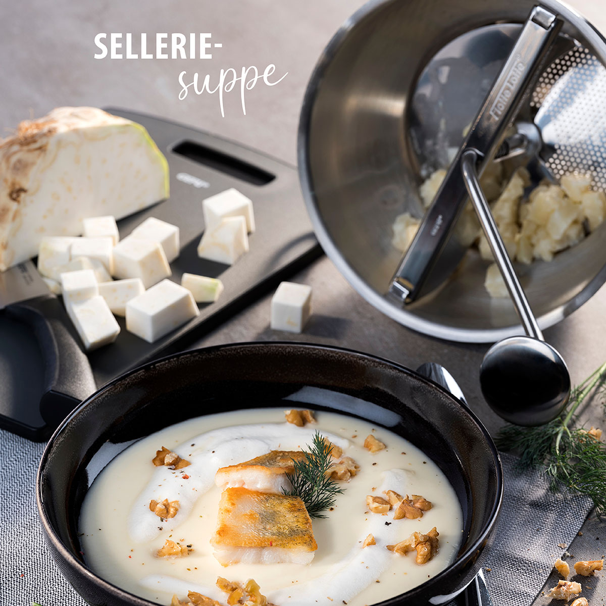 Cremige Selleriesuppe mit Fischfilet und Walnüssen Ein Teller cremige Selleriesuppe mit gebratenem Fischfilet und Walnüssen, daneben frisch gewürfelter Sellerie und eine Passiermühle auf einem Tisch.