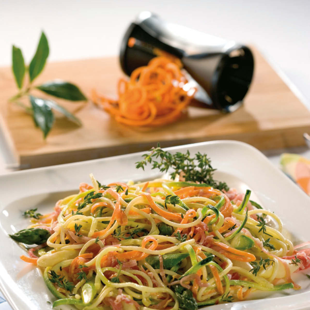 Leckere Gemüsenudeln mit Karottenspiralen und Kräutern Ein Teller mit frischen Gemüsenudeln, Karottenspiralen und Kräutern im Vordergrund, mit einem Spiralschneider und Karottenresten im Hintergrund
