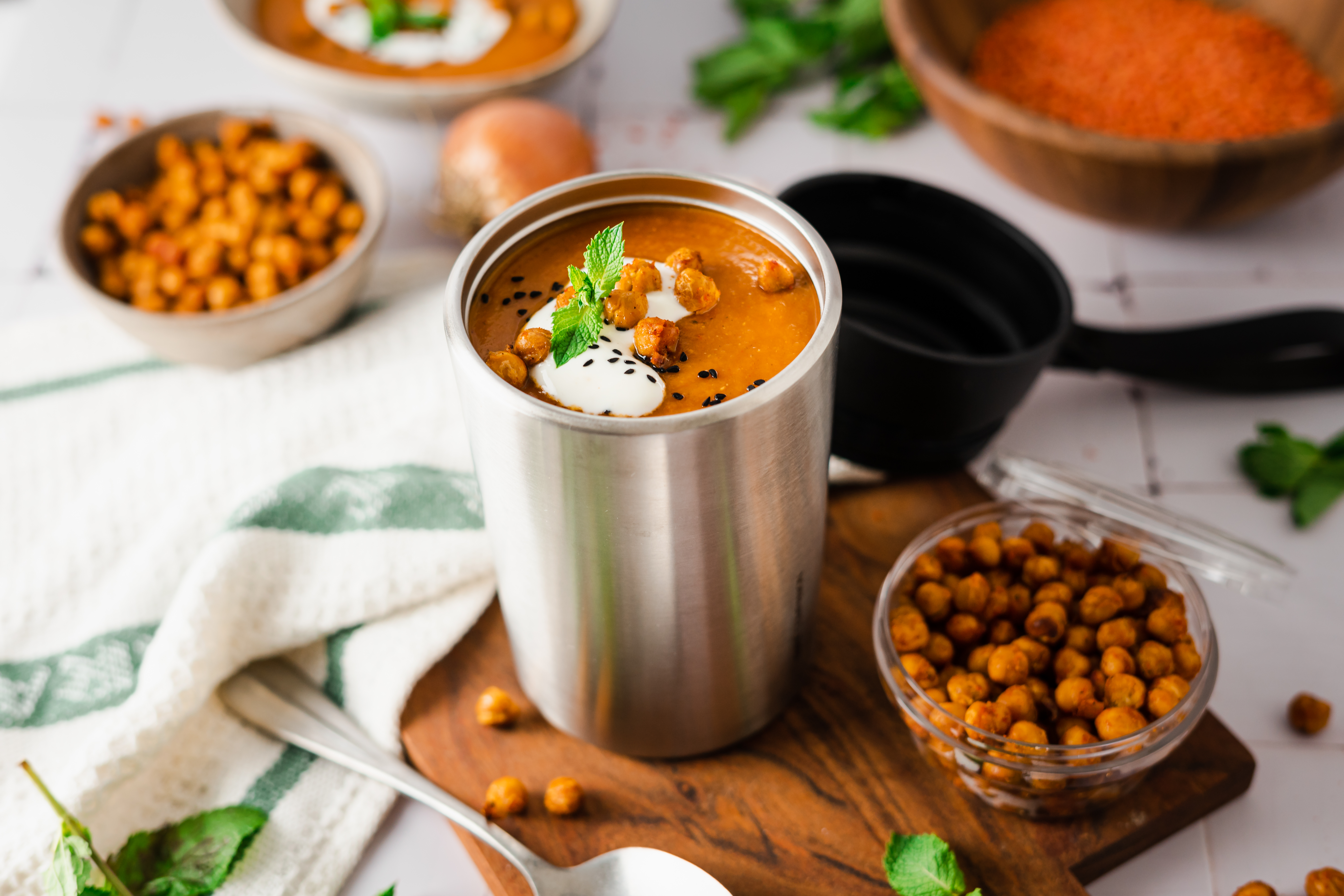 Türkische Linsensuppe mit gerösteten Kichererbsen  Ein wärmeisolierter Becher gefüllt mit cremiger Linsensuppe, garniert mit gerösteten Kichererbsen, frischer Minze und einem Klecks Joghurt, auf einem Holzbrett serviert.