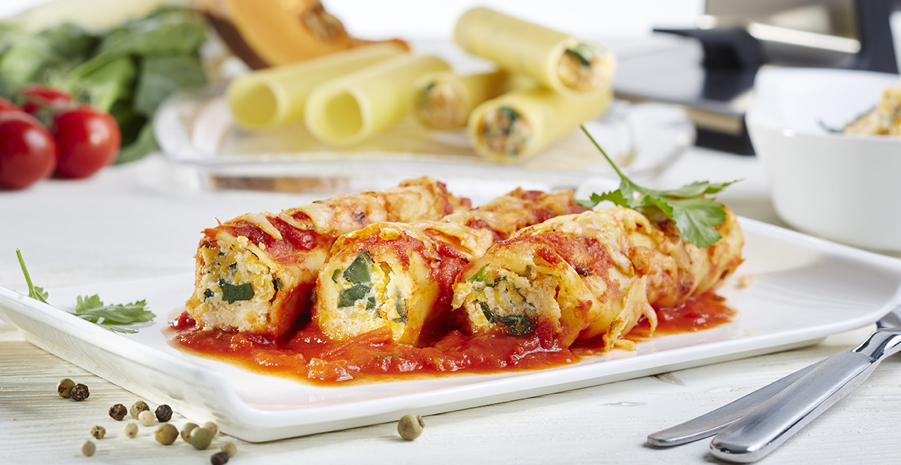 Cannelloni mit Kürbis-Mangold-Füllung Frische, gefüllte Cannelloni serviert mit Tomatensauce und Käse auf einem weißen Teller, im Hintergrund Zutaten wie Tomaten und Pasta-Rollen