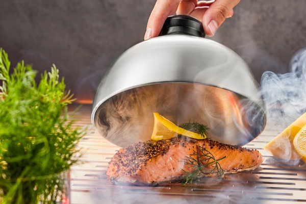 Ein Lachsfilet, frisch geräuchert und mit Kräutern garniert, wird unter einer silbernen Glocke präsentiert.