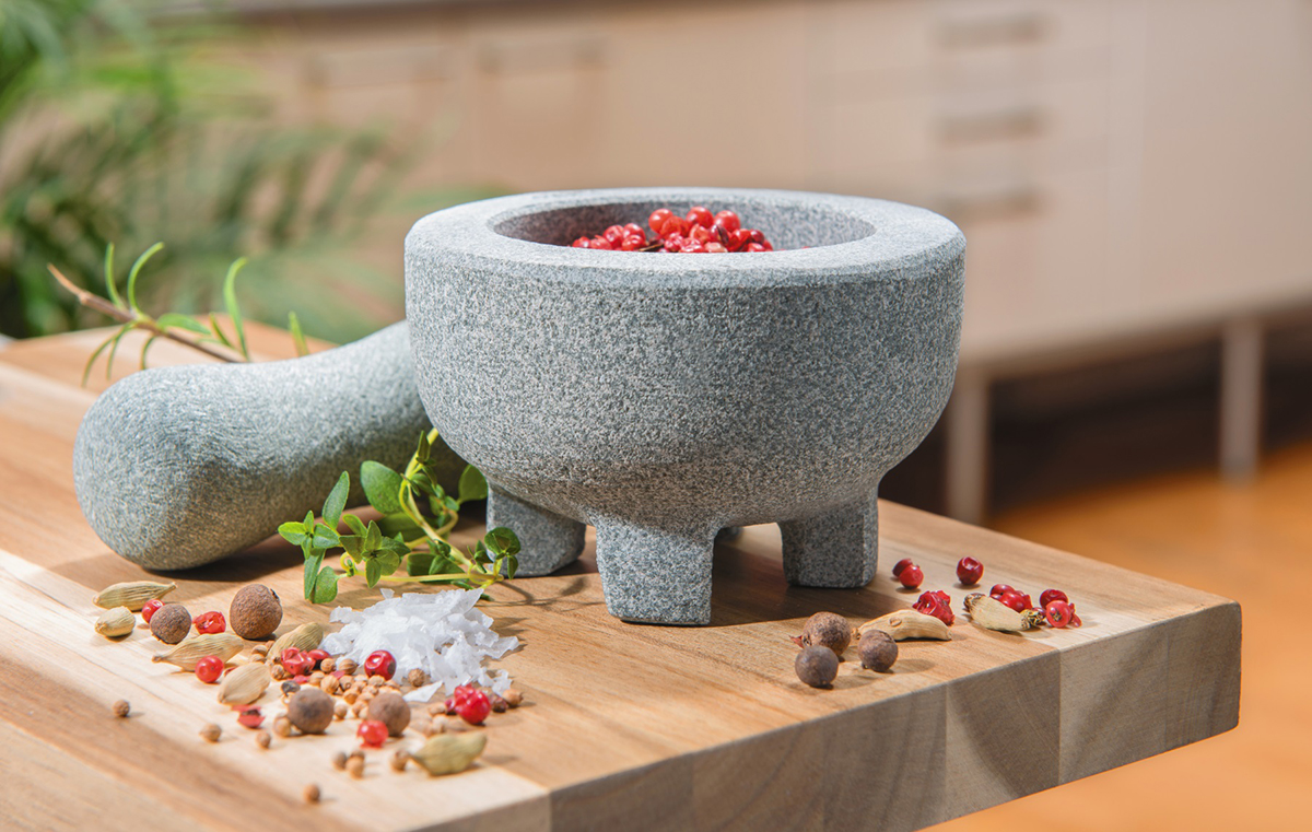 Granitmörser mit Stößel und Gewürzen auf Holztisch in moderner Küche Grauer Granitmörser mit Stößel und verschiedenen Gewürzen wie Pfeffer und Salz auf einem Holztisch in einer modernen Küche