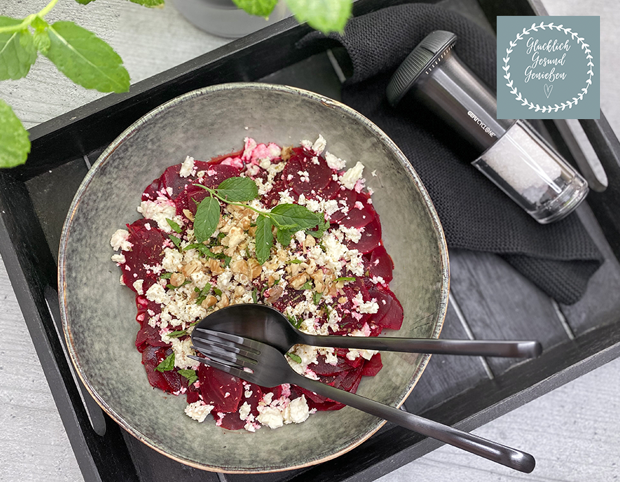 Rotebeete Carpaccio Ein Teller mit dünn geschnittenem Rote-Bete-Carpaccio, garniert mit zerbröckeltem Feta, gehackten Walnüssen und frischen Kräutern, serviert auf einem schwarzen Tablett mit Besteck.