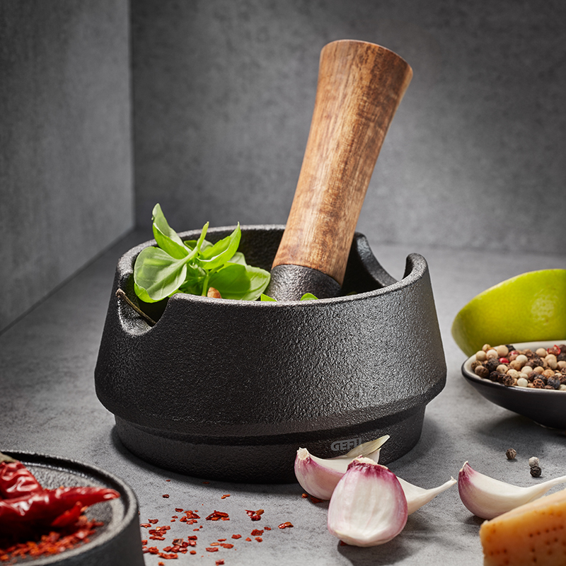 Hochwertiger Mörser aus Gusseisen mit Stößel und frischen Zutaten Nahaufnahme eines schwarzen Gusseisenmörsers mit Holzstößel, umgeben von frischen Kräutern, Knoblauch und Gewürzen auf einer Steinoberfläche.
