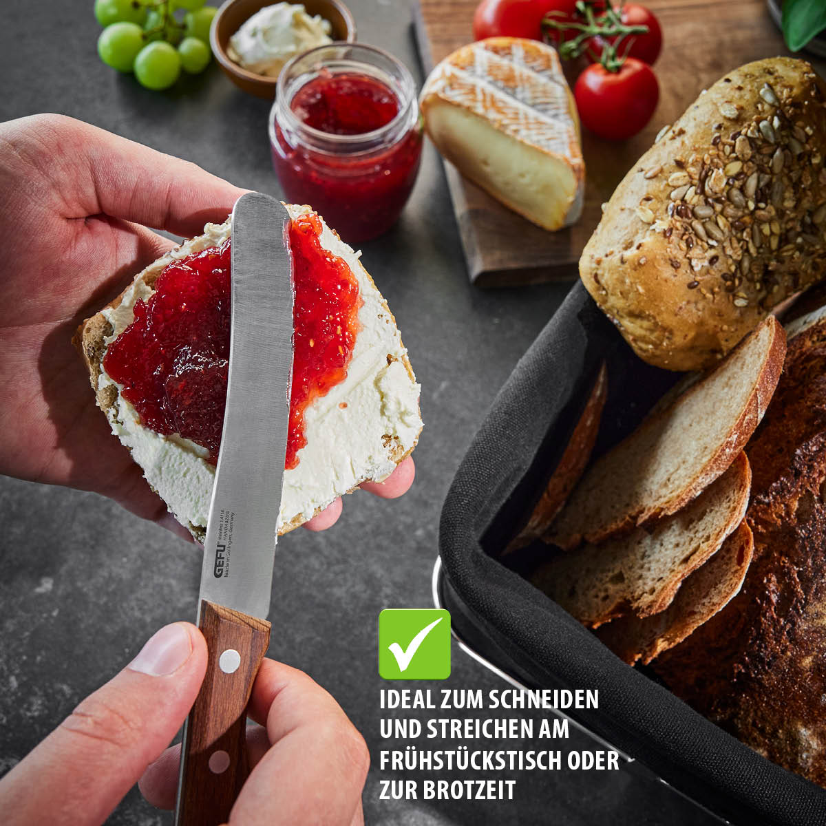 Hand hält Brötchen mit Frischkäse und Marmelade, umgeben von Brot und Käse