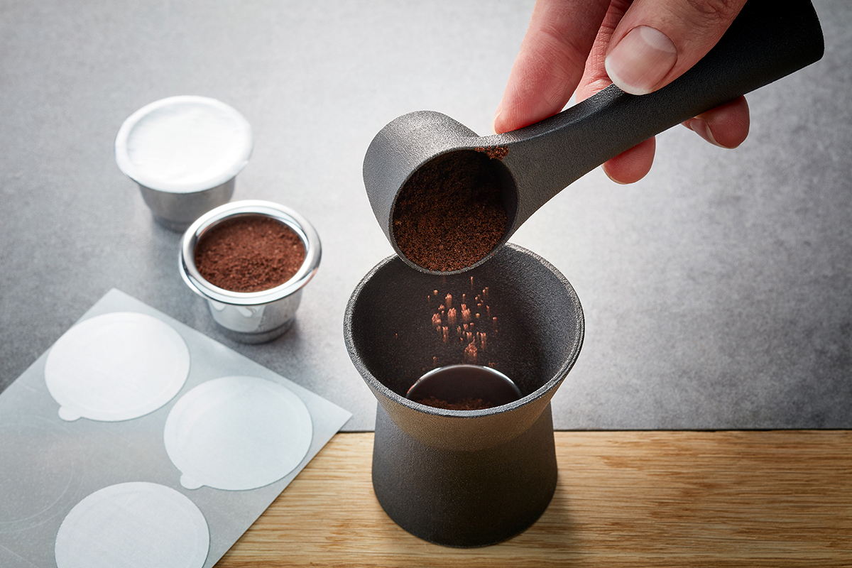 Kaffeepulver wird in eine Kapsel gefüllt - DIY Kaffeezubereitung Nahaufnahme von Kaffeepulver, das mit einem Löffel in eine leere Kaffeekapsel gefüllt wird. Im Hintergrund sind vorbereitete Kapseln und Versiegelungen zu sehen.