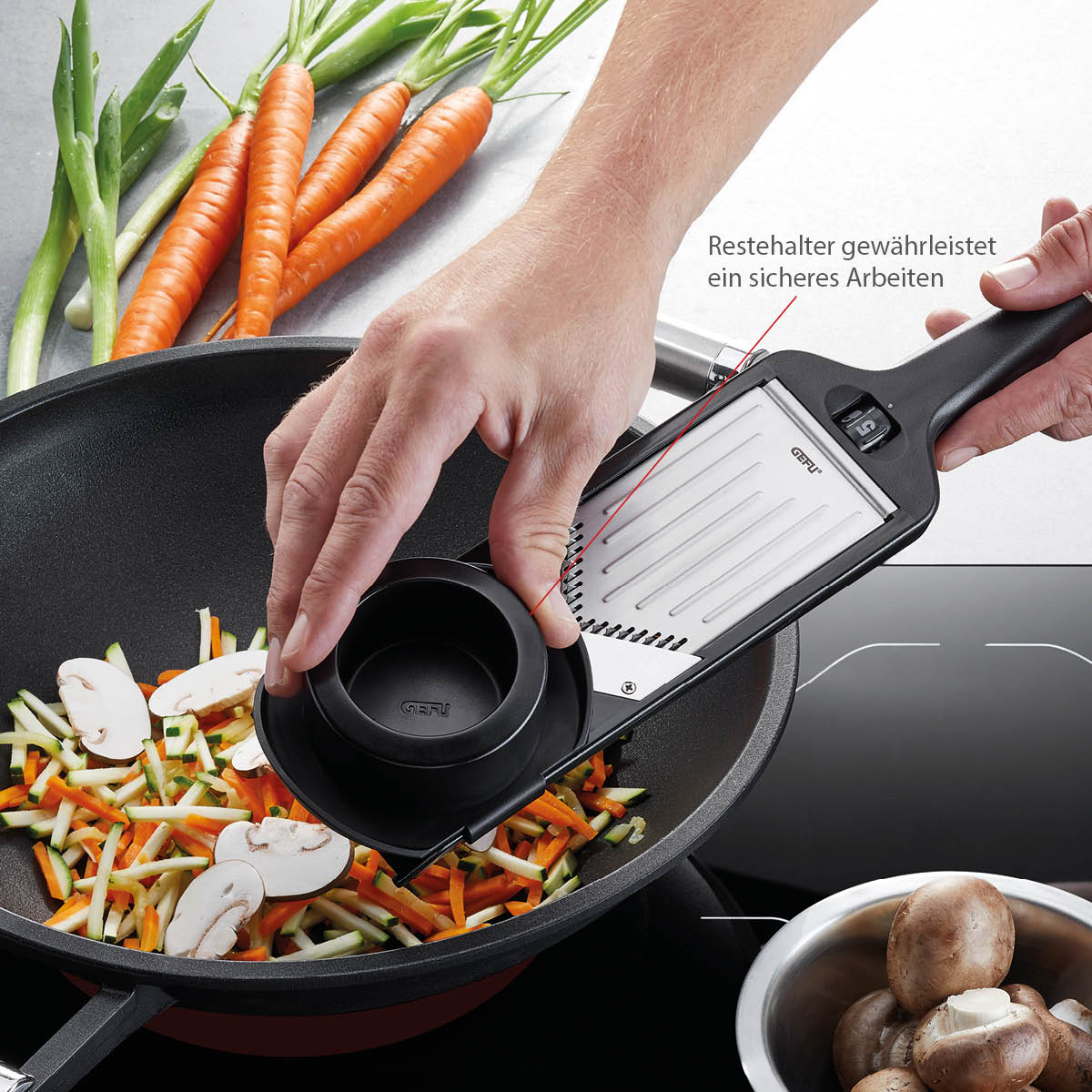Person verwendet einen Gemüsehobel mit Sicherheitshalter, um Karotten und Zucchini in einer Pfanne zu schneiden, frische Pilze und Karotten im Hintergrund