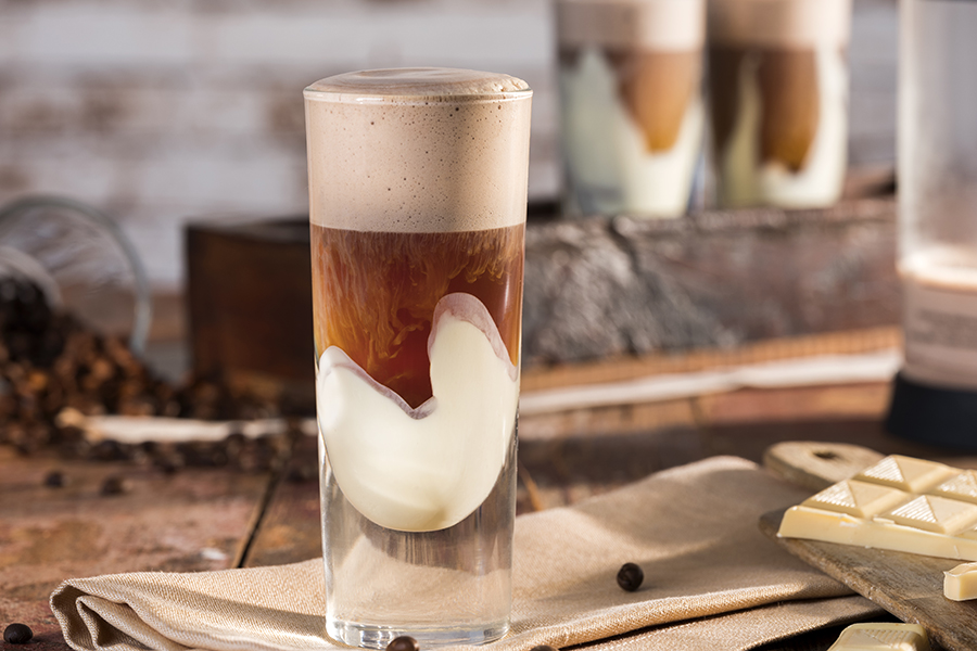 Caffè con cioccolato Ein Glas Latte Macchiato mit Sahne und Schokoladennote, auf einem Tisch mit Kaffeeschoten und weißer Schokolade.